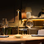 Lampe sans fil Solo dorée sur une table de restaurant, entre deux verres à vin