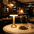 Lampe sans fil Fugue dorée allumée sur une table en travertin dans un bar à vin parisien